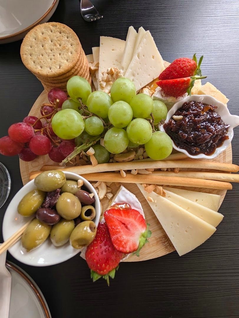 A round wooden board of sliced cheese, crackers, grapes, strawberries, olives, breadsticks and a dish of chutney